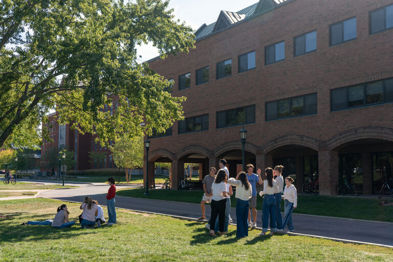 A group of young people gather on a grassy area near a brick building, with some sitting and others standing in conversation under a tree.